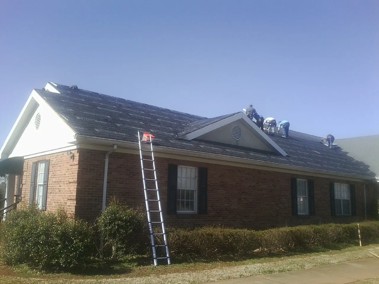 Crew tearing shingles off a brick ranch roof with a ladder set — Metal Roof Repair work by our crew in Puryear