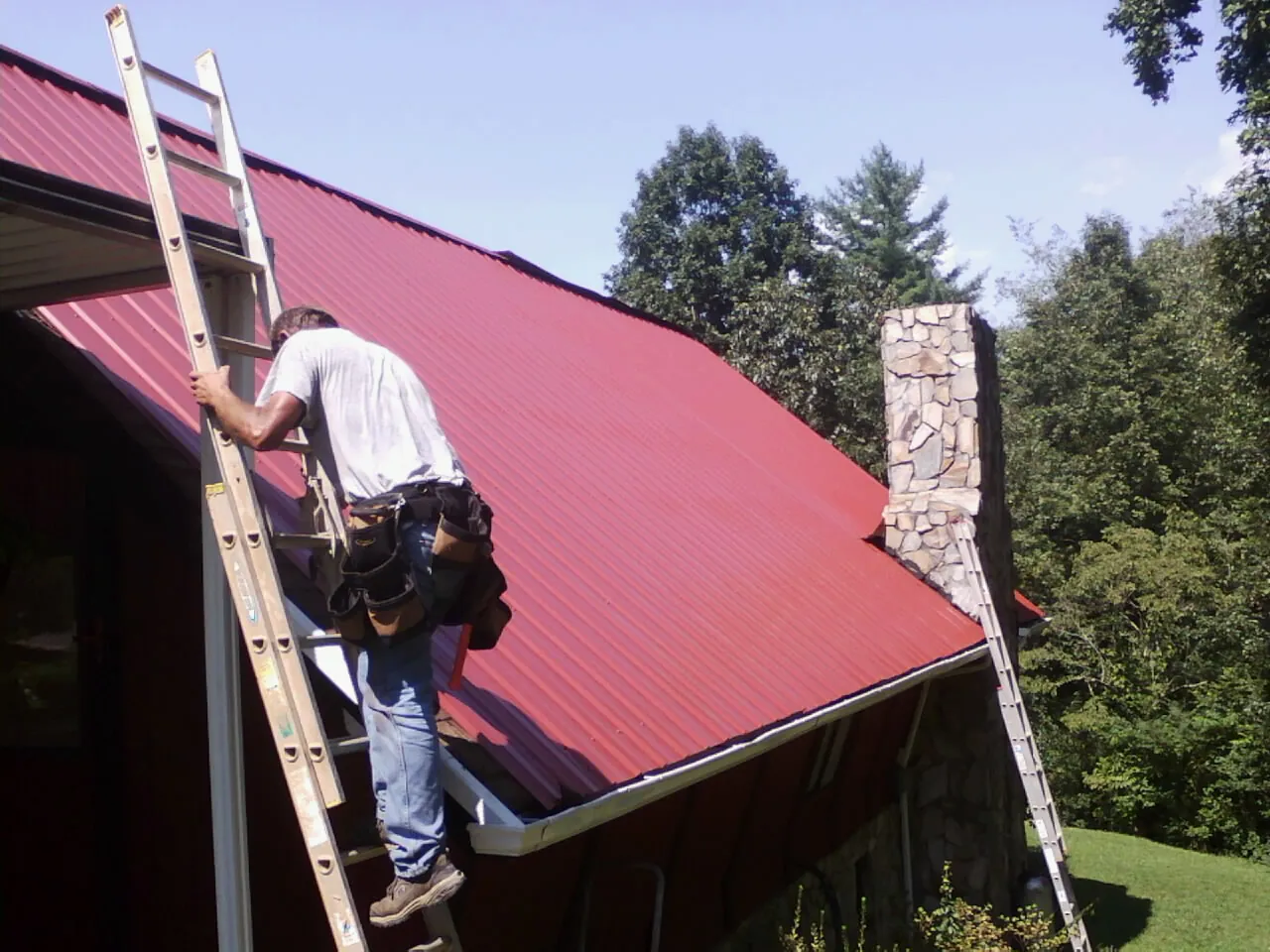 Roofer on a ladder beside a red metal roof and stone chimney — Soffit & Fascia Repair by Roofing Brothers in Puryear